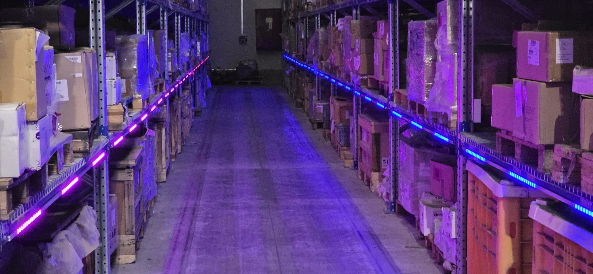 Long dim warehouse aisle with multi-color shelf-edge LED strips glowing into the distance
